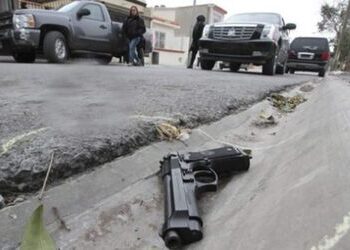A gun lies in the foreground in a gutter while a number of black cars and people go past in the background