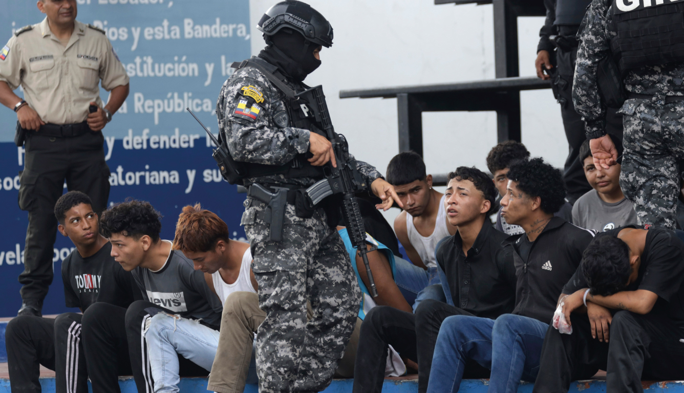 An Ecuadorian soldier stands in front of a line of seated, detained men.